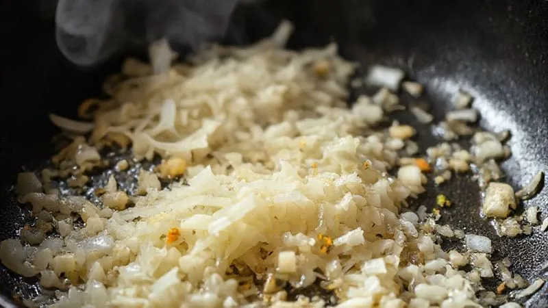 A close-up, photorealistic image of finely chopped onions, garlic, and ginger sizzling gently in a hot pan, turning golden brown, with steam rising. The scene suggests a meticulous cooking process, cinematic lighting, sharp focus on the ingredients, depth of field, 8K hyper-detailed, shot on a high-end DSLR.