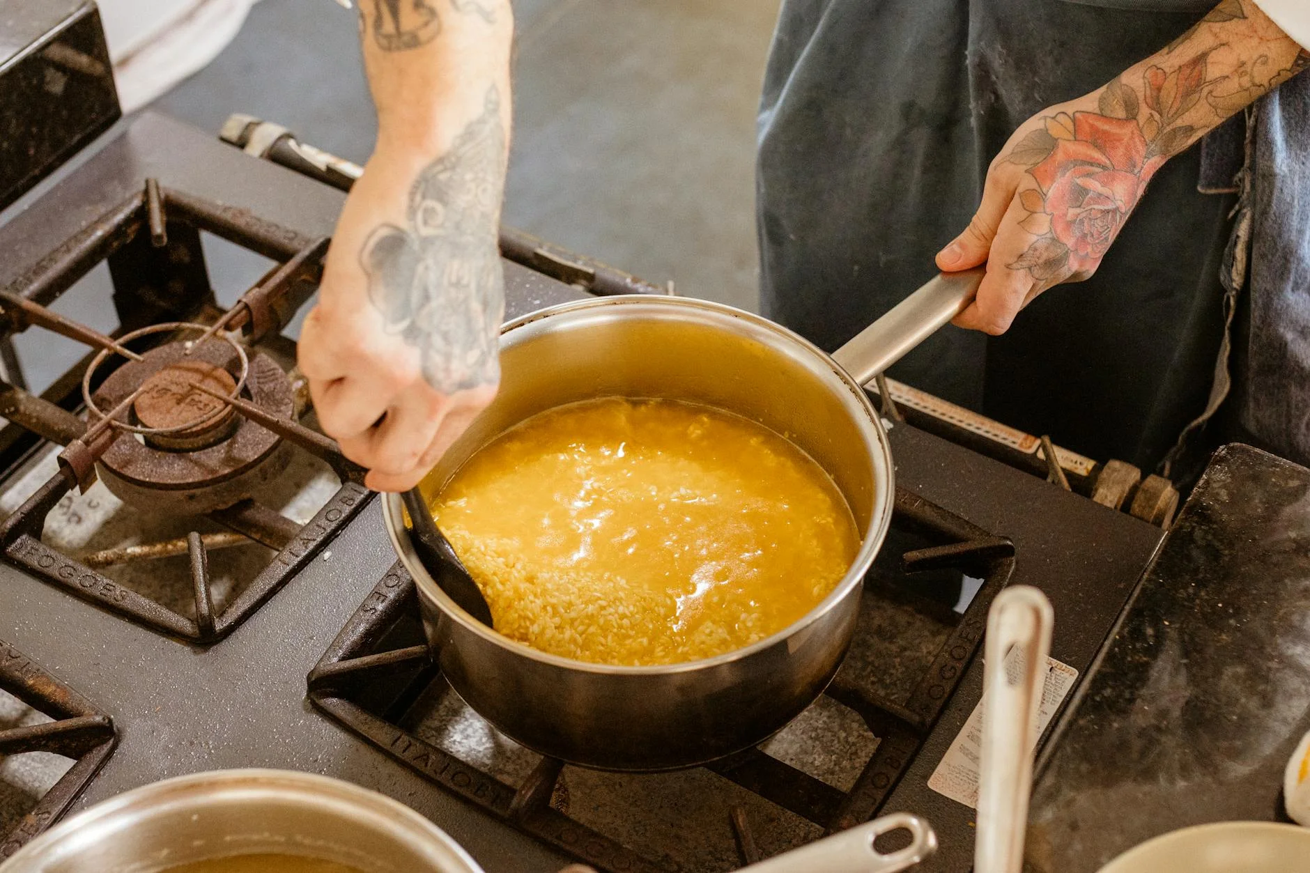 Tattooed chef stirring pasta on a stove while cooking in a kitchen. — Foto: Jonathan Borba / Pexels