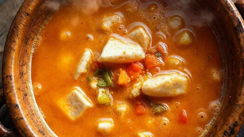A photorealistic overhead shot of a Moqueca Baiana simmering gently in a traditional clay pot, with the rich, golden-orange broth bubbling softly around chunks of white fish and colorful vegetables. The surface of the broth is visibly creamy and glistening with palm oil, and steam gently rises, creating an inviting, warm atmosphere. professional photography, 8K, cinematic lighting, sharp focus, depth of field, shot on a high-end DSLR.
